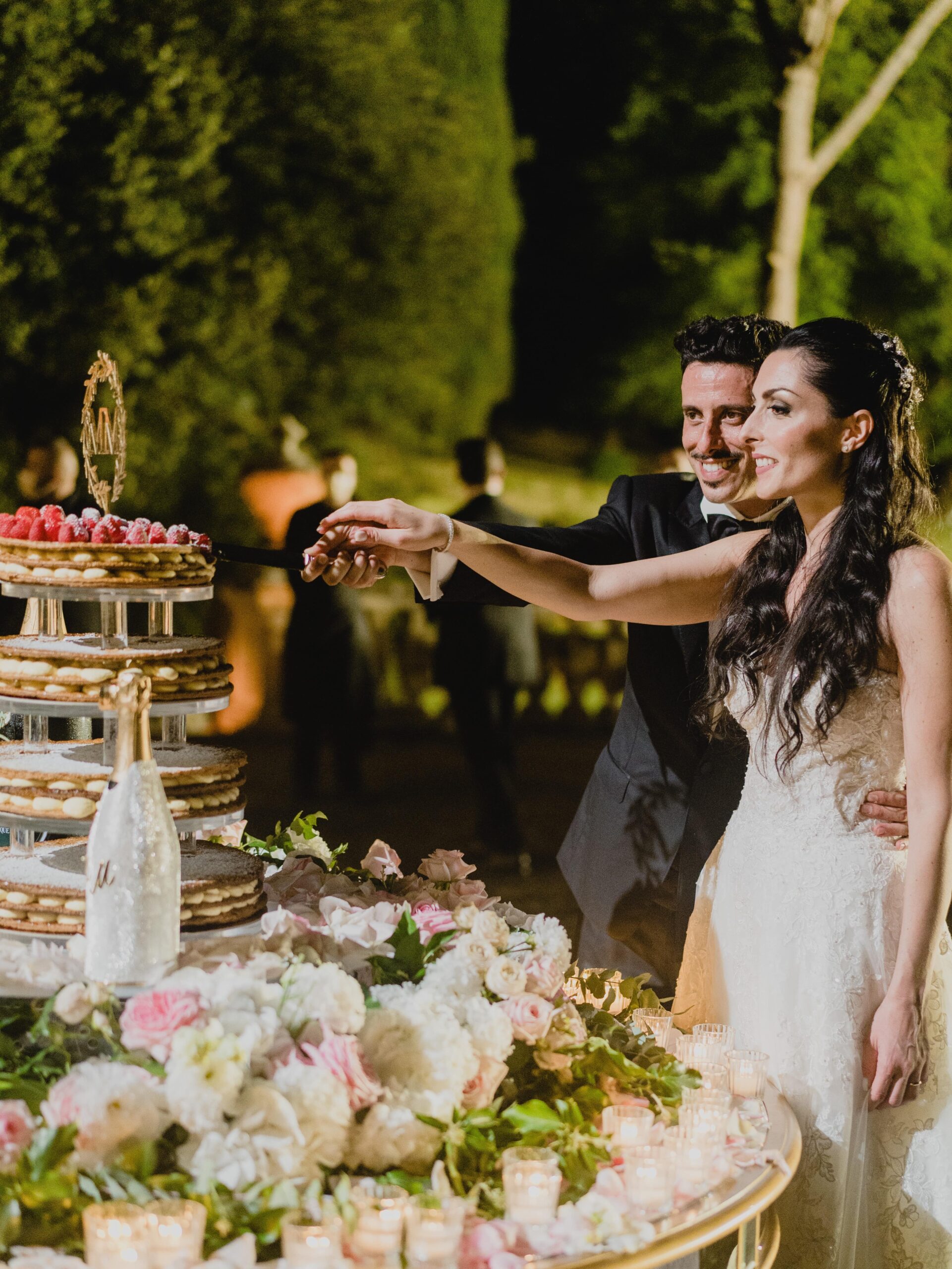 Bride and groom cutting their wedding cake at an outdoor reception, with a decorated champagne bottle featuring a personalized silver cover placed beside the cake