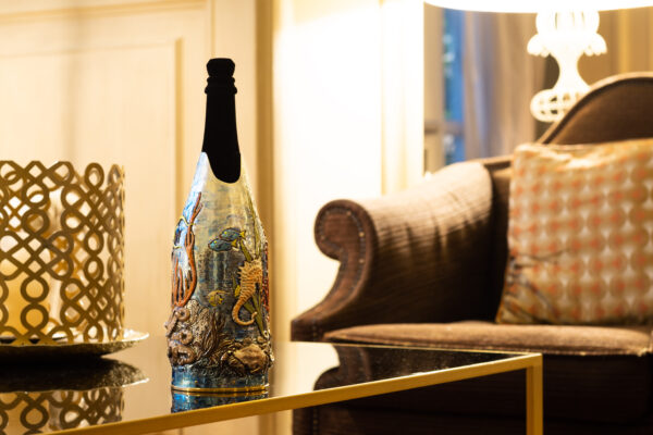 Champagne Cover AQUARIUM. Champagne bottle with 'Acquarium' cover, a colorful, hand-painted metal artwork depicting an underwater scene with corals, fish, and a seahorse
