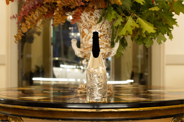 The champagne bottle is dressed with the 'Ariosto' cover, a silver creation inspired by the epic tales of Ludovico Ariosto. The cover features a finely sculpted battle scene between a knight and a dragon, evoking the imagery of Orlando Furioso. Positioned on a richly decorated inlaid table, with a grand vase and autumnal foliage in the background, the setting enhances the sense of art, history and luxury.