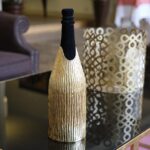 The champagne bottle covered with the 'Canne' cover, a handcrafted gold sheath with vertical reed-like textures, placed on a reflective black and gold table. In the background, a decorative gold lantern with an intricate geometric pattern complements the luxurious aesthetic of the scene.