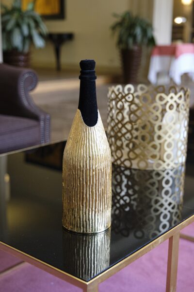 The champagne bottle covered with the 'Canne' cover, a handcrafted gold sheath with vertical reed-like textures, placed on a reflective black and gold table. In the background, a decorative gold lantern with an intricate geometric pattern complements the luxurious aesthetic of the scene.