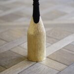 A champagne bottle wrapped in the 'Canne' cover, a handcrafted gold piece with a vertical textured pattern reminiscent of natural reed stems. The cover’s organic design contrasts elegantly with the geometric wooden floor, highlighting the fusion of nature-inspired artistry and refined luxury.