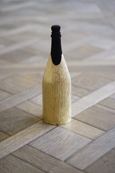A champagne bottle wrapped in the 'Canne' cover, a handcrafted gold piece with a vertical textured pattern reminiscent of natural reed stems. The cover’s organic design contrasts elegantly with the geometric wooden floor, highlighting the fusion of nature-inspired artistry and refined luxury.