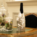 A champagne bottle embellished with the 'Rushmore' cover, a handcrafted silver sculpture wrapping the lower part of the bottle. The design features a three-dimensional face emerging from the metal, resembling a modern artistic tribute to monumental sculptures. Positioned on an elegant wooden desk with glass jars filled with colorful candies, the scene evokes a sense of refined craftsmanship and luxurious indulgence.