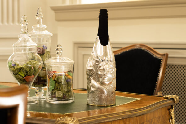 A champagne bottle embellished with the 'Rushmore' cover, a handcrafted silver sculpture wrapping the lower part of the bottle. The design features a three-dimensional face emerging from the metal, resembling a modern artistic tribute to monumental sculptures. Positioned on an elegant wooden desk with glass jars filled with colorful candies, the scene evokes a sense of refined craftsmanship and luxurious indulgence.
