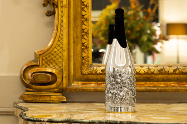 The champagne bottle adorned with the 'Battaglia di Cascina' cover, a silver artwork that transforms the bottle into a sculptural masterpiece. The cover depicts a dynamic battle scene in high relief, inspired by historical art. Placed on a marble surface in front of an ornate gold mirror, the setting enhances the sense of opulence and timeless craftsmanship.