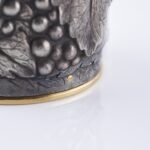 Close-up of a silver champagne bottle cover with detailed engraving of grape clusters and vine leaves, finished with a gold base trim
