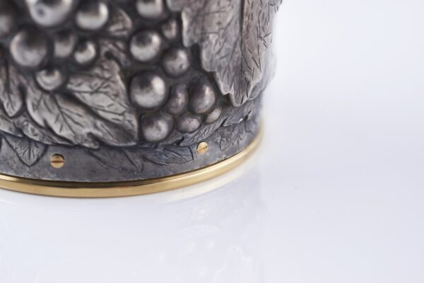 Close-up of a silver champagne bottle cover with detailed engraving of grape clusters and vine leaves, finished with a gold base trim