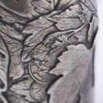 Close-up of a silver champagne bottle cover with detailed engraving of grape clusters and vine leaves, finished with a gold base trim