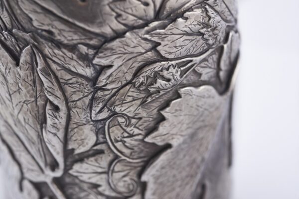 Close-up of a silver champagne bottle cover with detailed engraving of grape clusters and vine leaves, finished with a gold base trim
